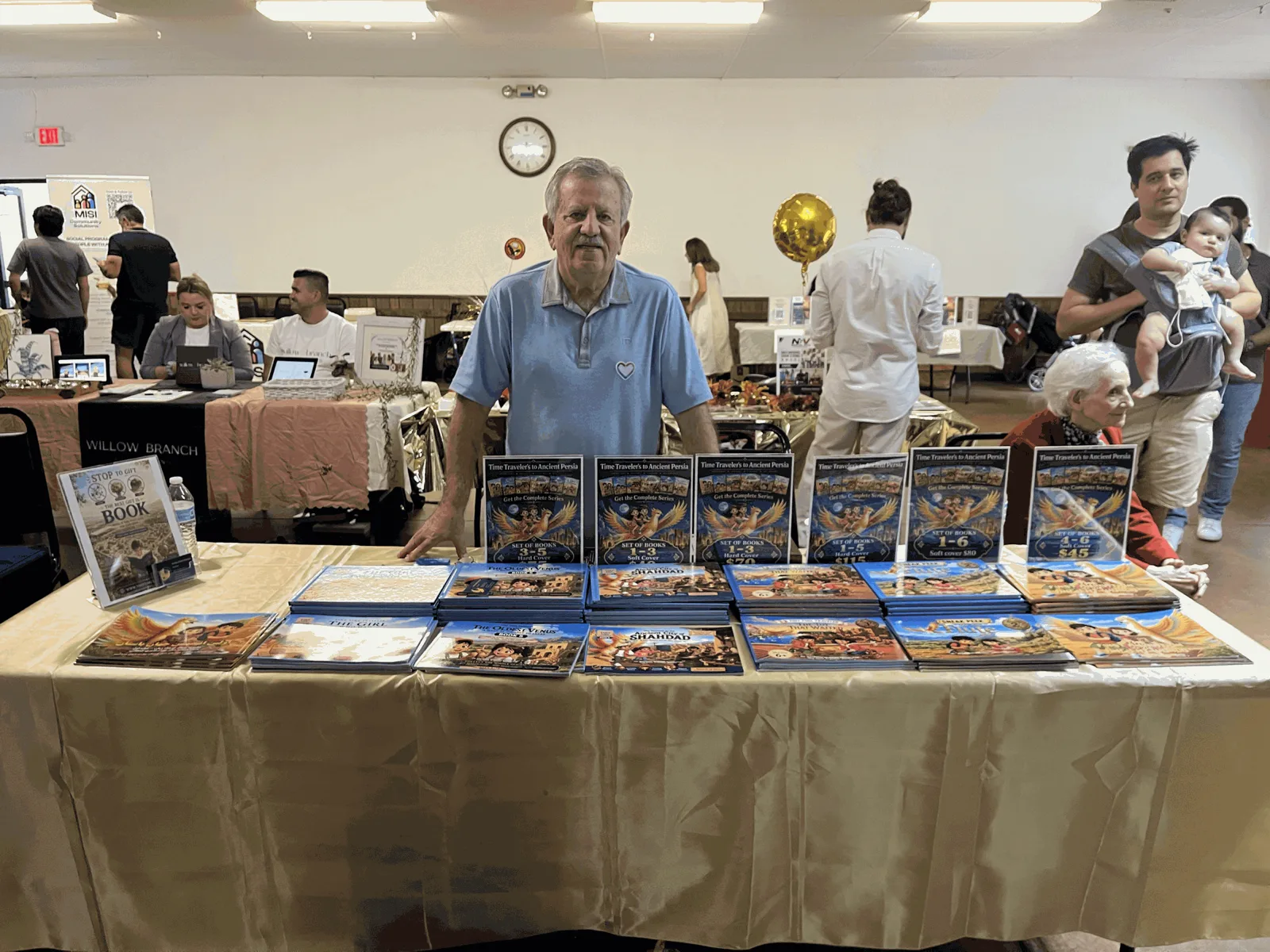 Dr. Bahram Ghadiri standing behind a long table of Tiny Time Traveler books at an indoor community fair, with set-price cards for the book bundles and a stack of the Persian edition.