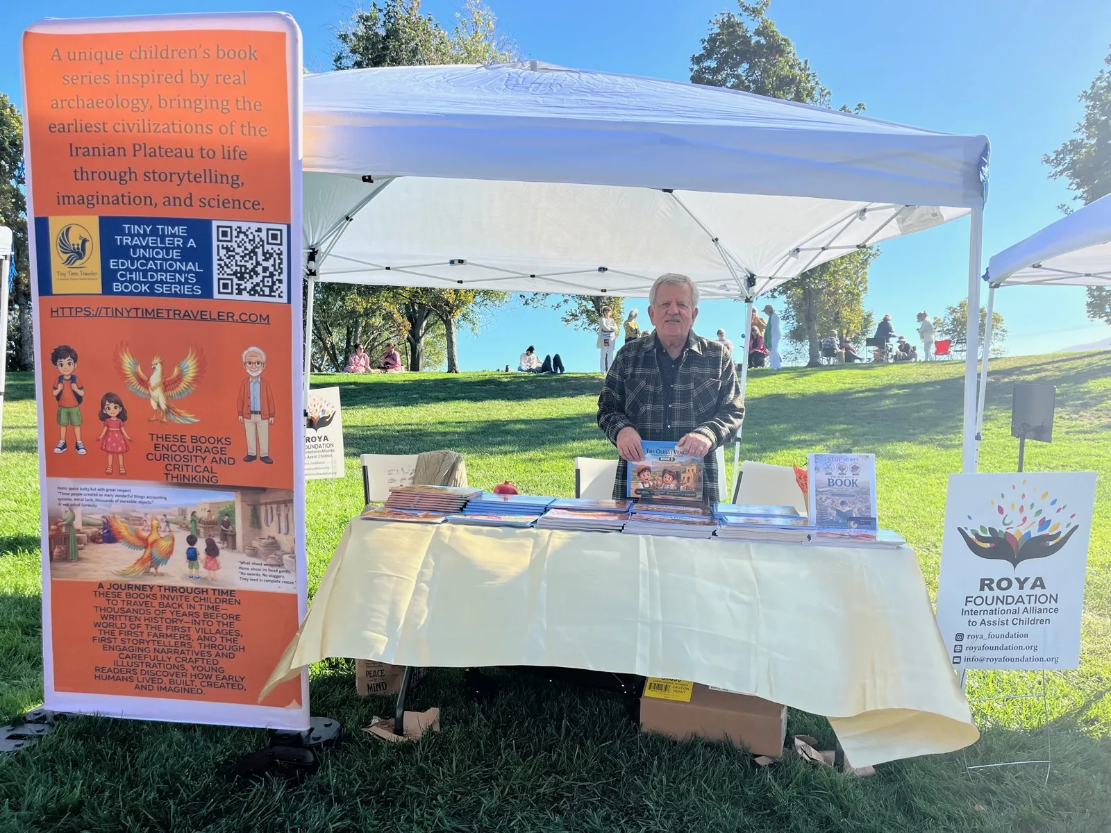 Dr. Bahram Ghadiri behind the Tiny Time Traveler table at an outdoor festival, holding a copy of The Oldest Venus, with the orange series banner and a stack of books.