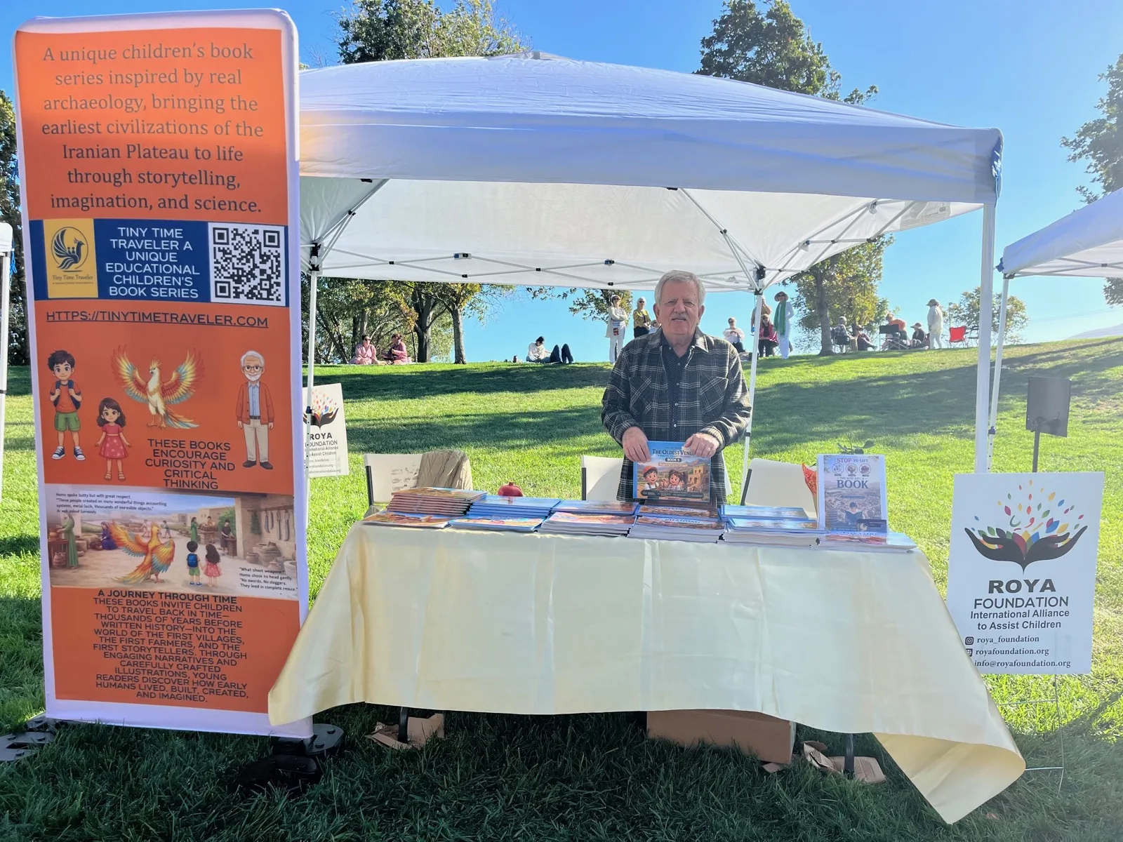Dr. Bahram Ghadiri holding a copy of The Oldest Venus behind the Tiny Time Traveler table under a white canopy in San Ramon Central Park, with the series banner beside him.