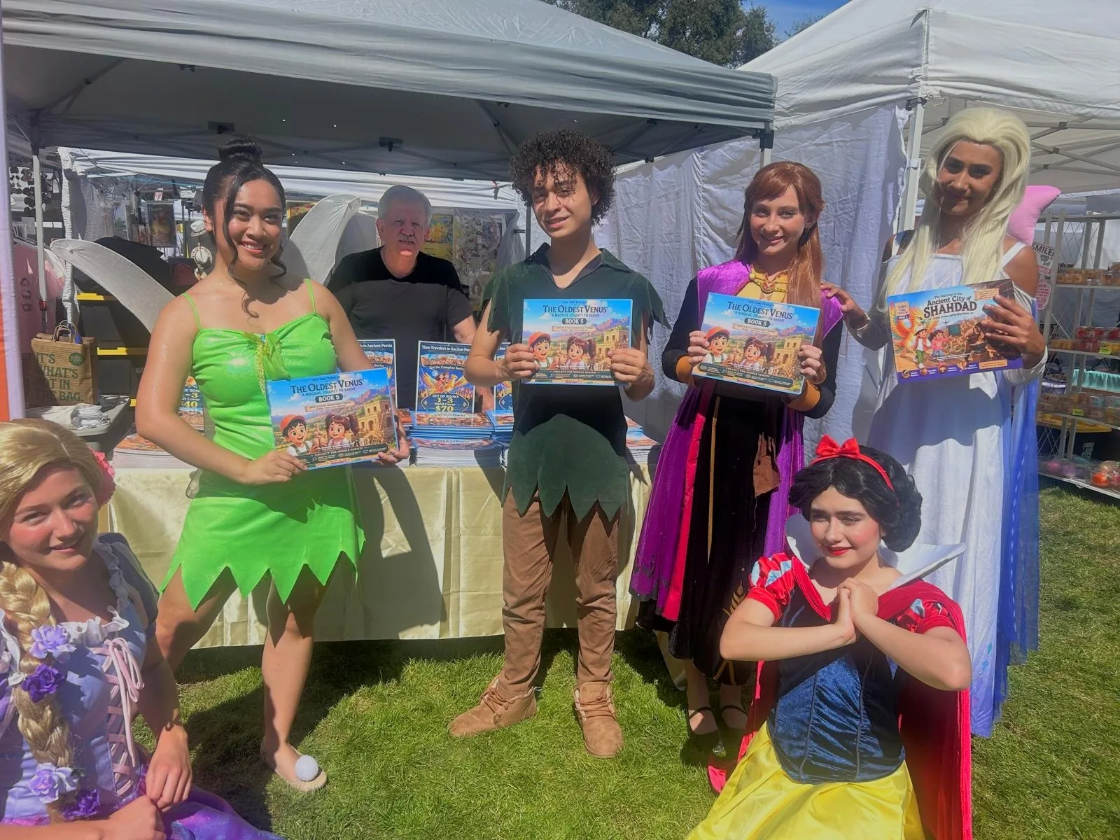 The six costumed characters at the Tiny Time Traveler table, holding copies of The Oldest Venus and Shahdad, with Dr. Bahram Ghadiri behind them.