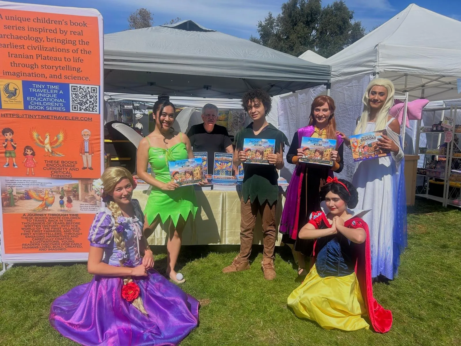 Rapunzel, Tinker Bell, Peter Pan, Anna, Elsa and Snow White in costume at the Tiny Time Traveler booth, holding copies of The Oldest Venus and The Journey to the Ancient City of Shahdad, with the orange Tiny Time Traveler banner at left and Dr. Bahram Ghadiri behind the book table.