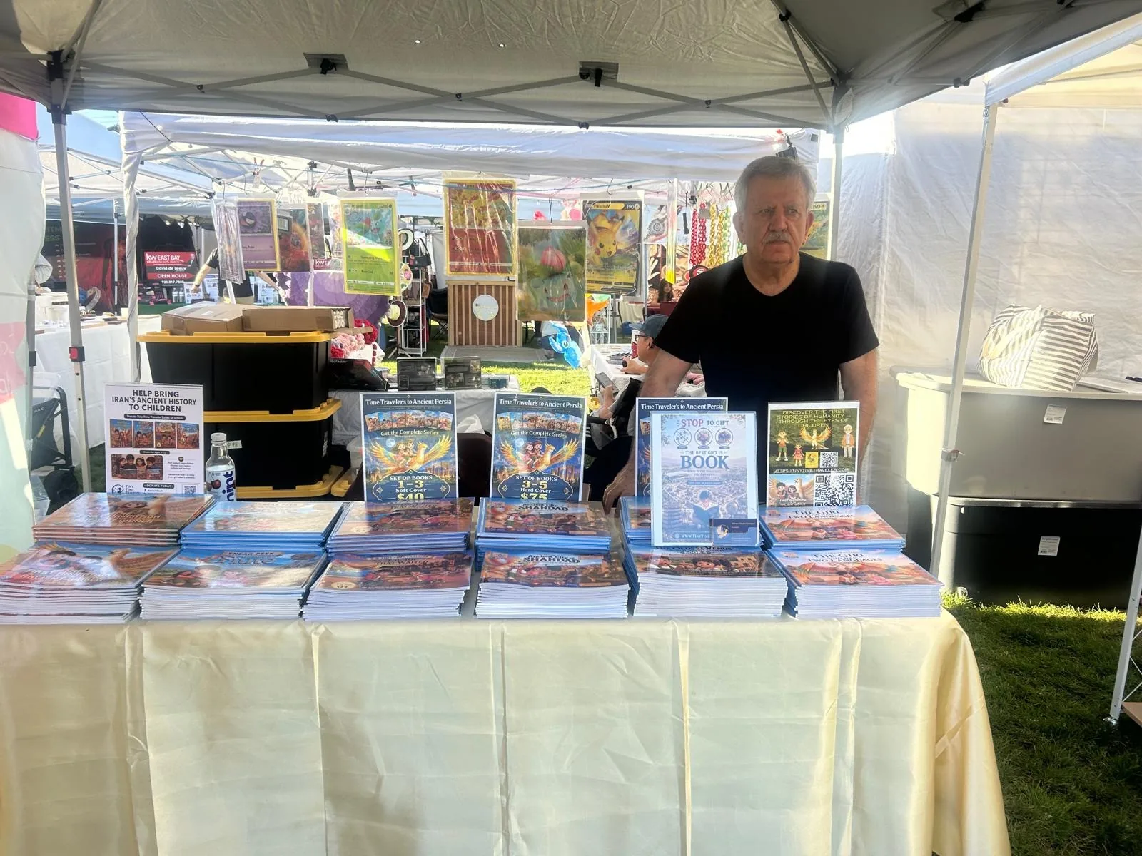 The Tiny Time Traveler table on Friday evening: stacks of all six books on a cream tablecloth, set-price cards for Books 1–3 and 3–5, and Dr. Bahram Ghadiri standing behind the table.